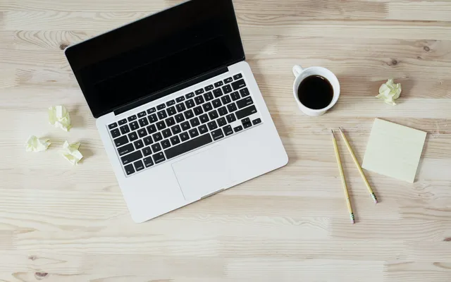 An open laptop and a cup of coffee on a light wooden desk, seen from above