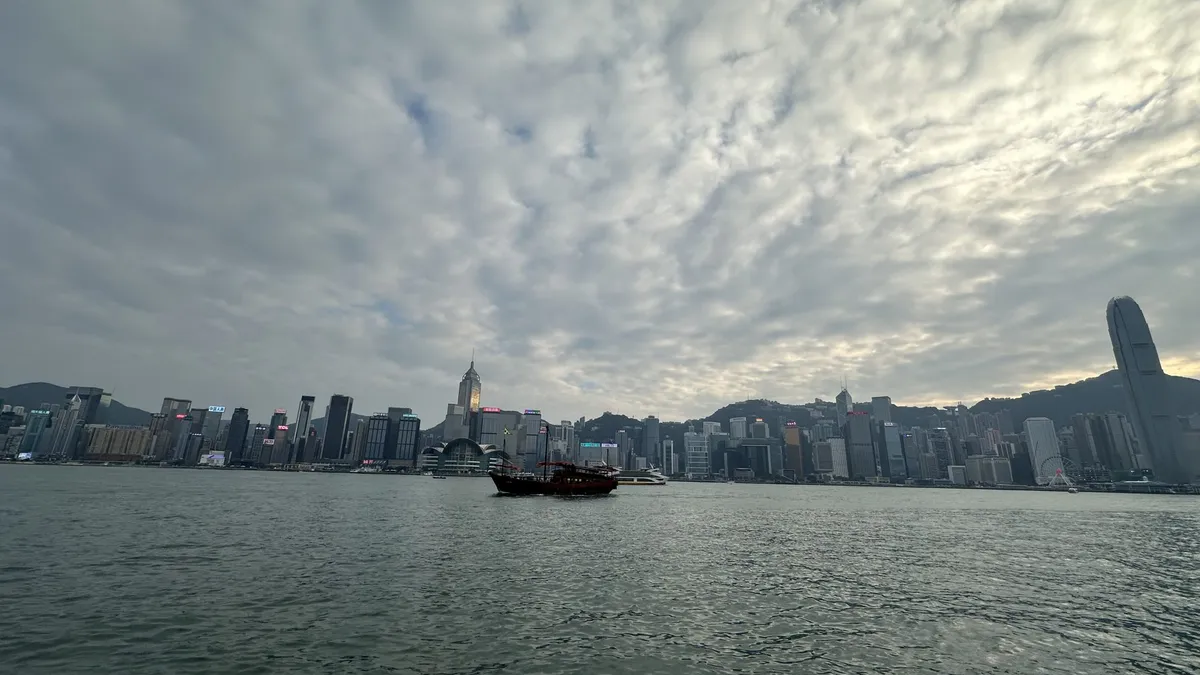 Victoria Harbour and the Hong Kong Island skyline under a wide cloudy sky, with a junk on the water