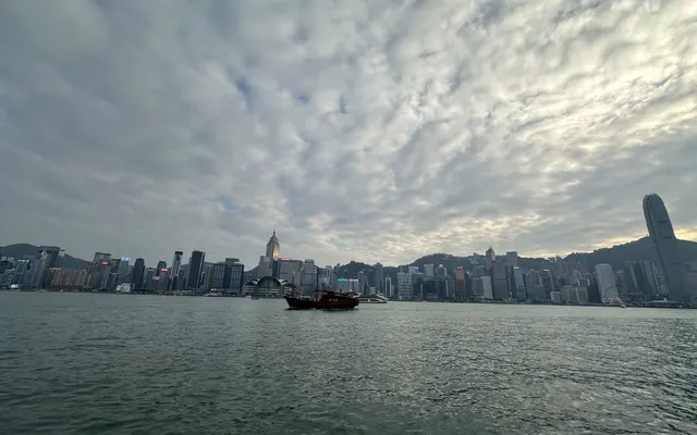 Victoria Harbour and the Hong Kong Island skyline under a wide cloudy sky, with a junk on the water