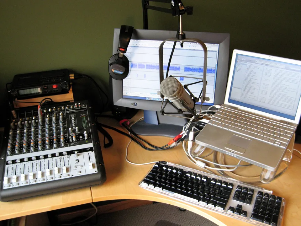 A home recording desk with a mixer, a hanging microphone, a monitor and a laptop