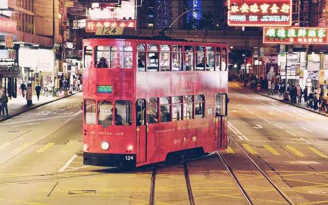 A red double-decker tram at night on a wide street lined with neon signs